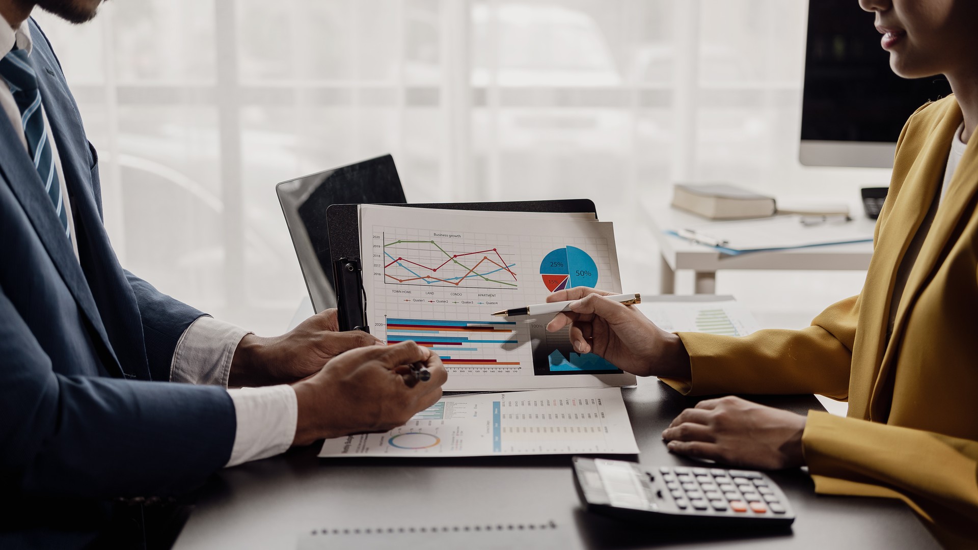 Two business partner colleagues discuss financial and company planning graph during budget meeting. Business partner uses tablet to analyze company financial budget report graph.